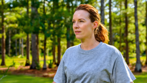 Woman in a gray t-shirt standing outdoors among tall pine trees on a sunny, humid Gulf Coast day