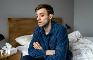 Tired man with flu symptoms in a navy plaid flannel shirt sitting on the edge of his bed with arms crossed, weighing flu treatment near Vancleave