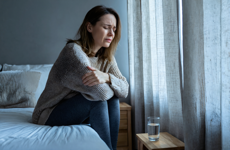 Woman with a migraine sitting on her bed in a dim room with the curtains drawn and a glass of water on the nightstand