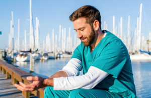 Man in teal scrubs looking at his inner forearm, where a TB skin test is placed, on a harbor boardwalk