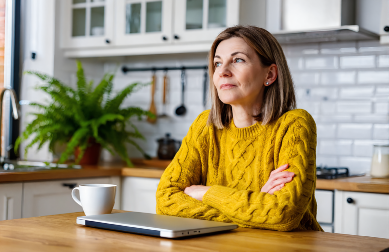 Woman in a yellow sweater at her kitchen table with arms folded, a closed laptop and coffee mug in front of her, weighing urgent care cost with insurance